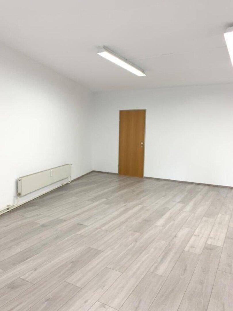 Leerer Raum mit hellem Laminatboden, weißen Wänden und einem Heizkörper unter dem Fenster.