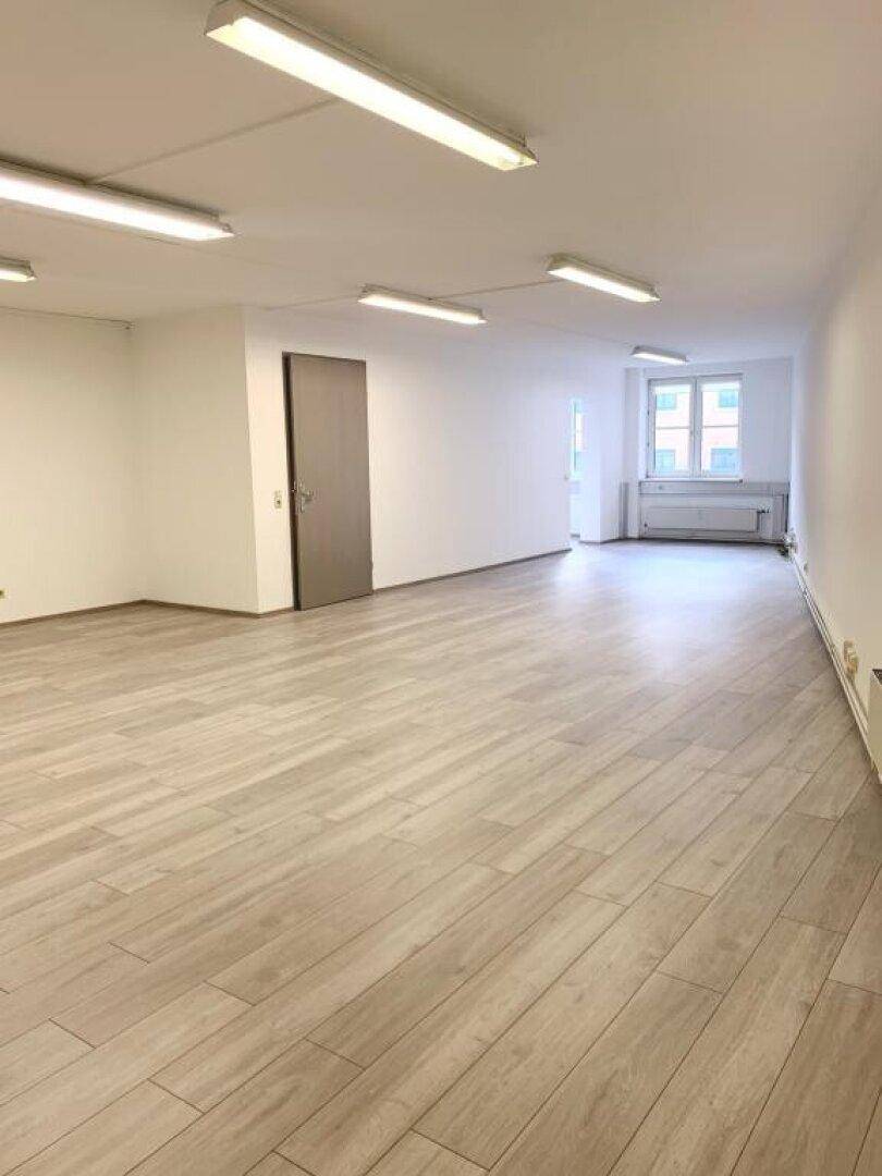 Heller, leerer Büroraum mit modernem Laminatboden und großen Fenstern für viel Tageslicht.