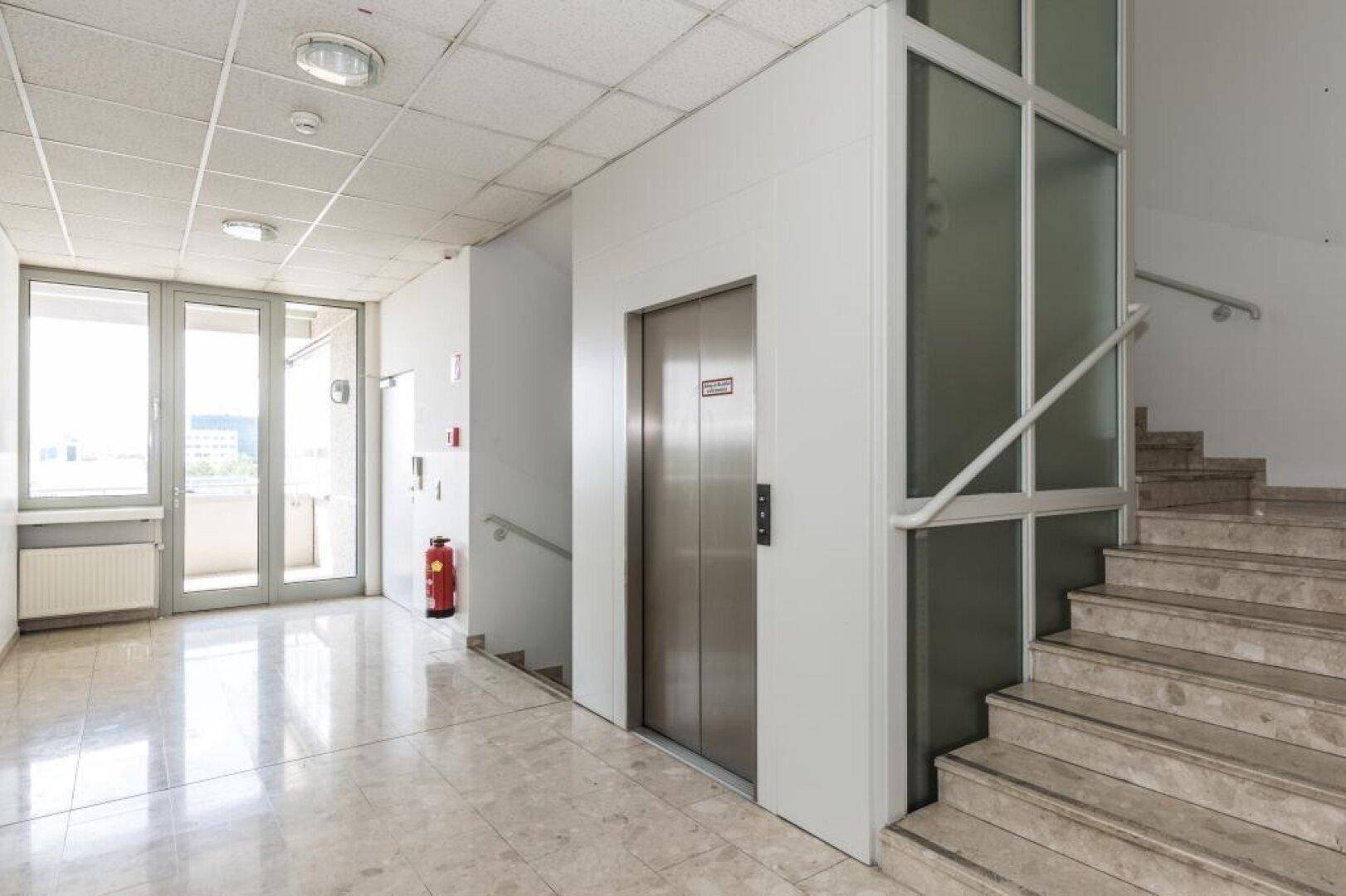 Heller Flur mit Marmorboden, Treppe und einem modernen Aufzug, der Zugang zu den Etagen bietet.