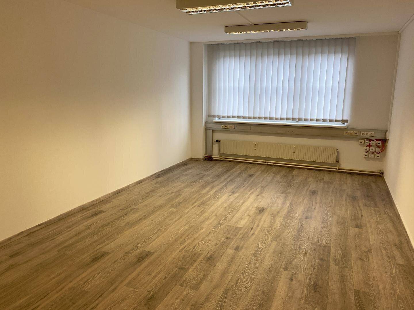 Geräumiger, leerer Büroraum mit Laminatboden und großem Fenster mit Jalousien.
