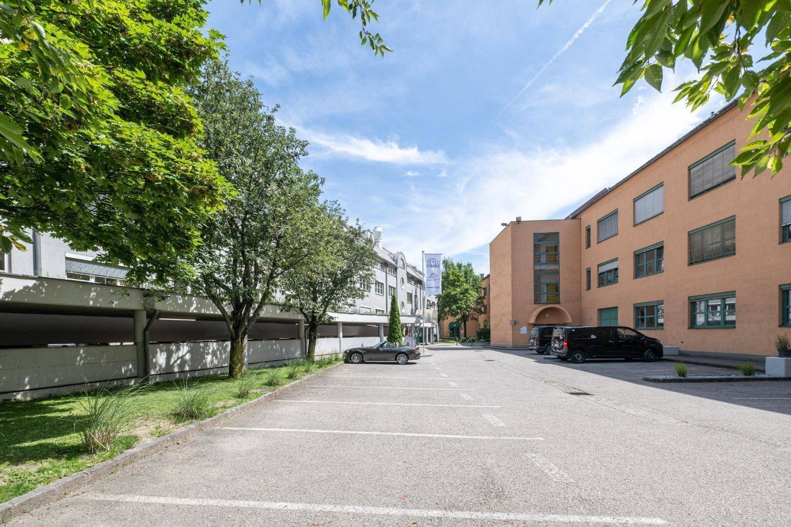 Außenansicht eines Parkplatzes mit umliegenden Gebäuden und viel Grün an einem sonnigen Tag.