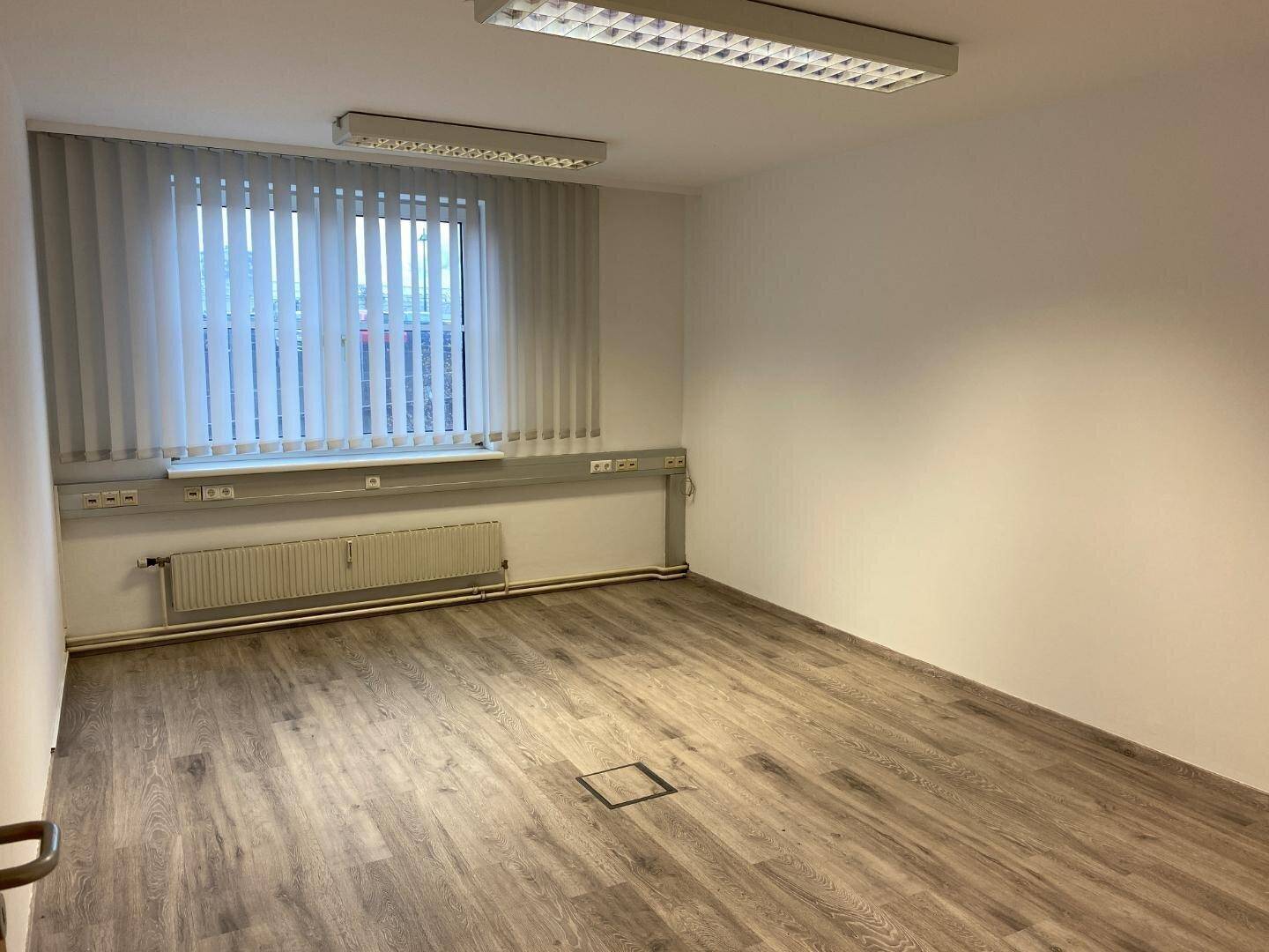 Leerer Büroraum mit Laminatboden, Fenster und Bodentank für flexible Arbeitsplatzgestaltung.