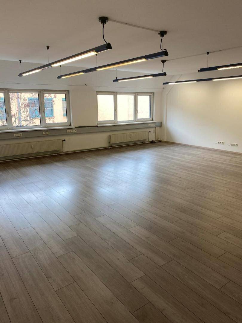 Leerer Büroraum mit vielen Fenstern, hellem Laminatboden und modernen, länglichen Deckenlampen.