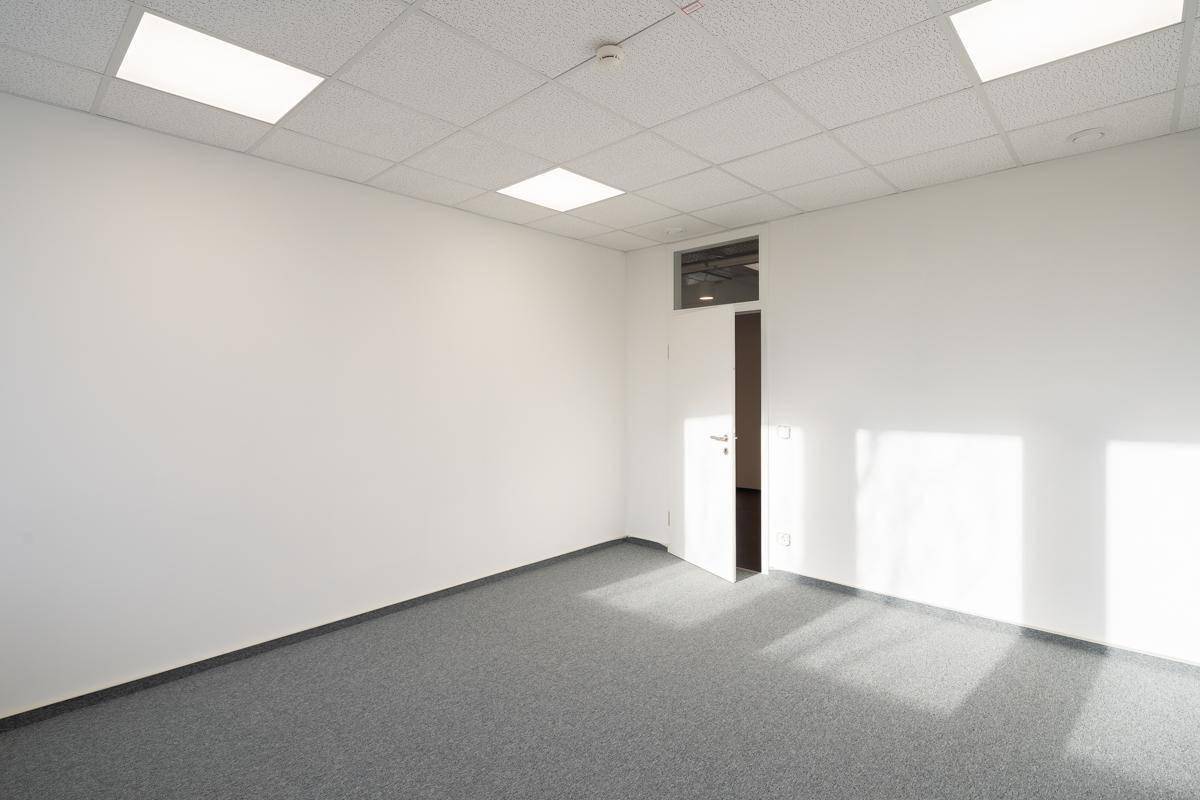 Leeres Büro mit weiß gestrichenen Wänden, grauem Teppichboden und einer offenen Tür zu einem weiteren Raum.