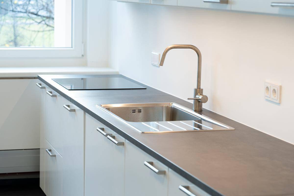 Moderne Küchenzeile mit Edelstahlspüle, Induktionskochfeld und weißen Unterschränken unter einem Fenster.