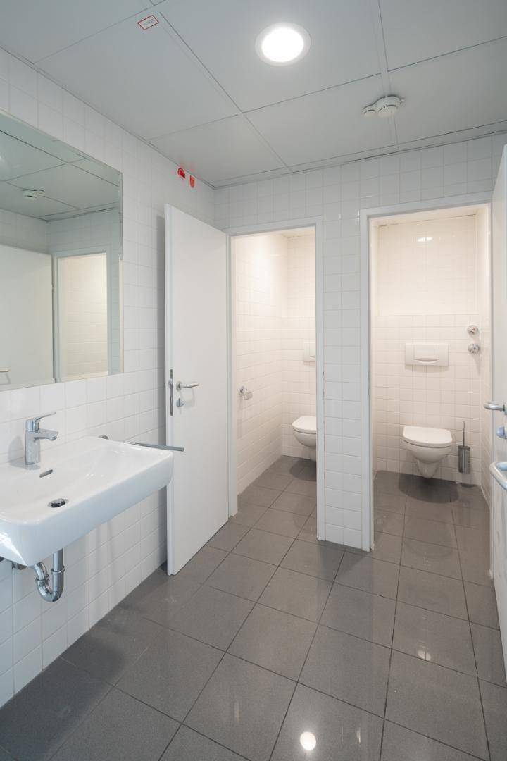 Moderner Sanitärbereich mit Waschbecken, Spiegel und zwei Toilettenkabinen, gefliest in Weiß und Grau.