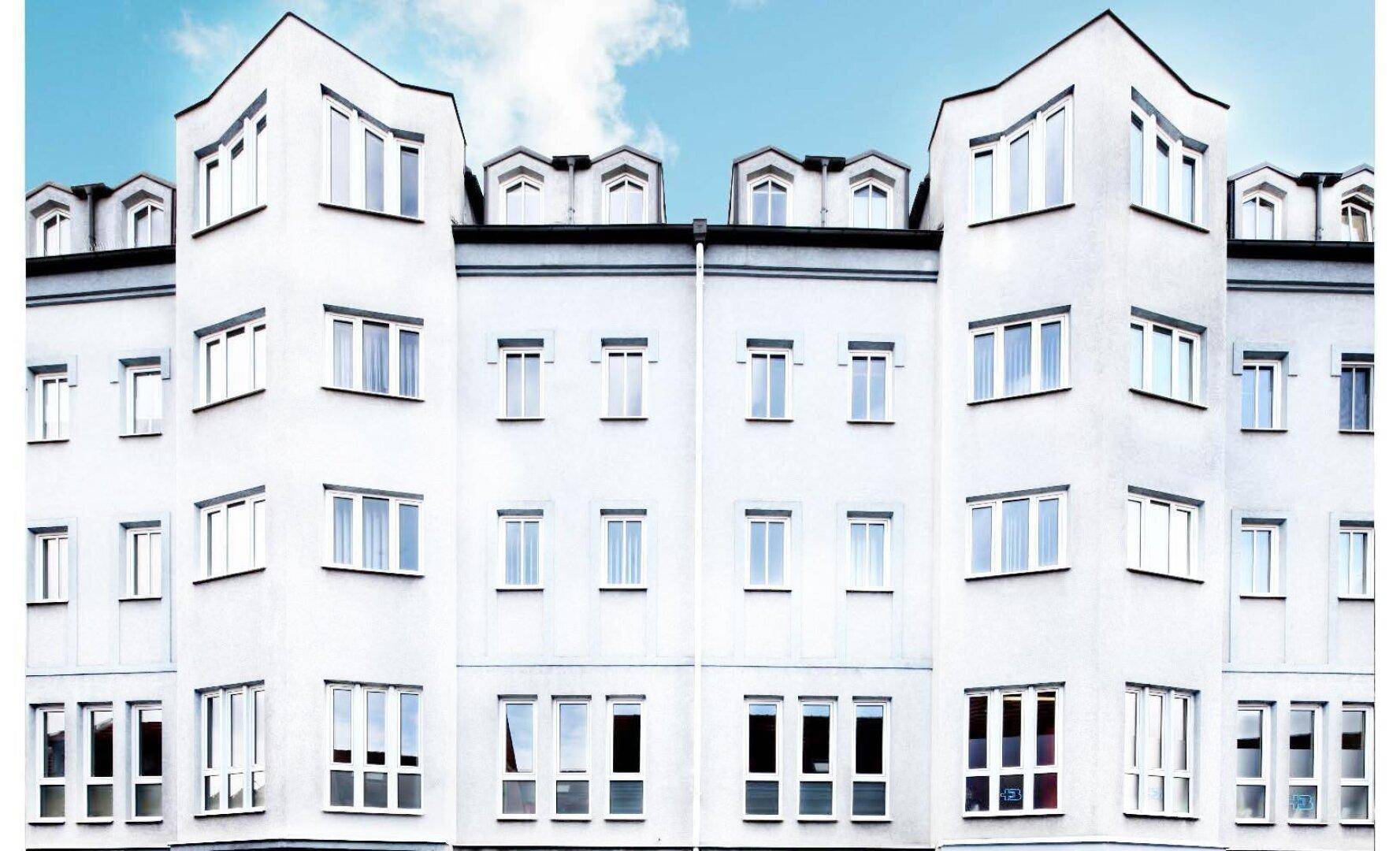Moderne weiße Fassade eines Mehrfamilienhauses mit vielen Fenstern und markanten Dachgauben unter blauem Himmel.