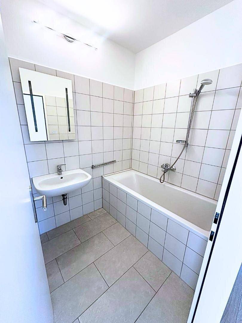 Funktionales Badezimmer mit Badewanne, Waschbecken, Haltegriffen und hellen Fliesen an Wänden und Boden.