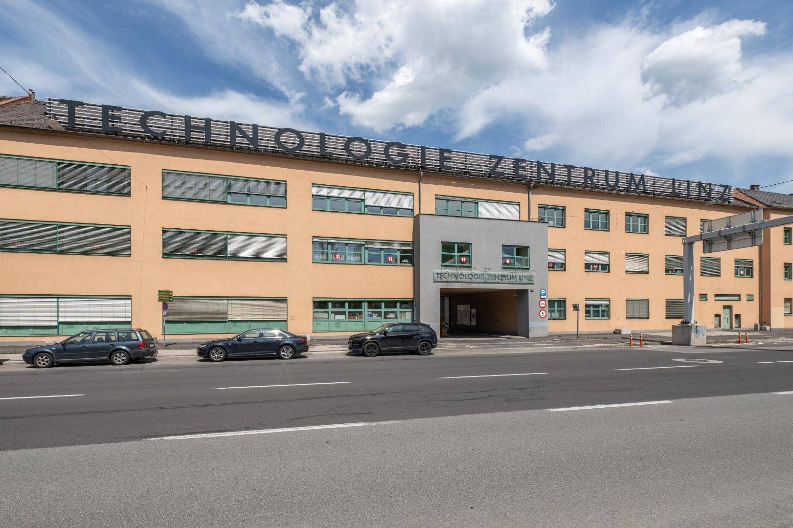 Außenansicht des Technologie Zentrums Linz mit gelber Fassade und geparkten Autos davor.