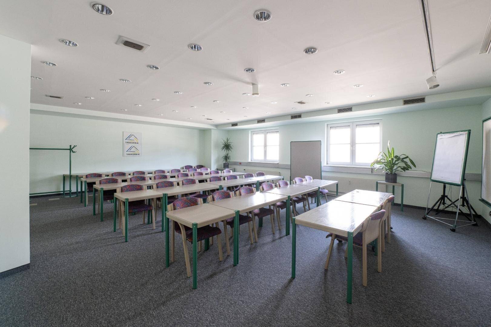 Großer Schulungsraum mit Reihen von Tischen und Stühlen, großen Fenstern und einem Whiteboard für Unterricht.
