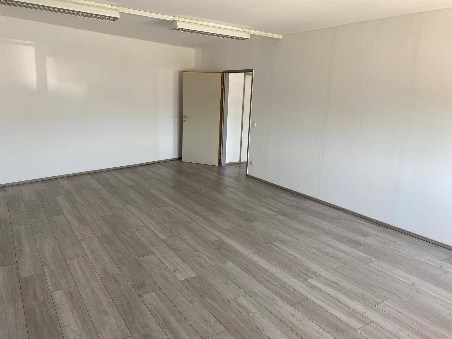 Geräumiger, leerer Büroraum mit hellem Laminatboden und weißen Wänden, der Zugang zu weiteren Räumen bietet.