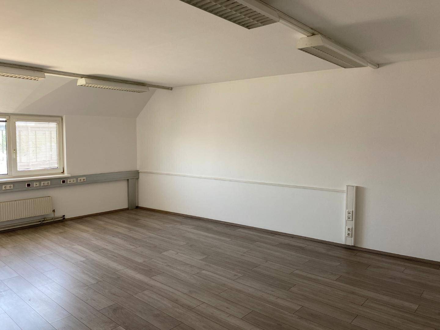 Heller Büroraum mit Laminatboden und weißen Wänden, ausgestattet mit Heizkörpern und Deckenbeleuchtung.