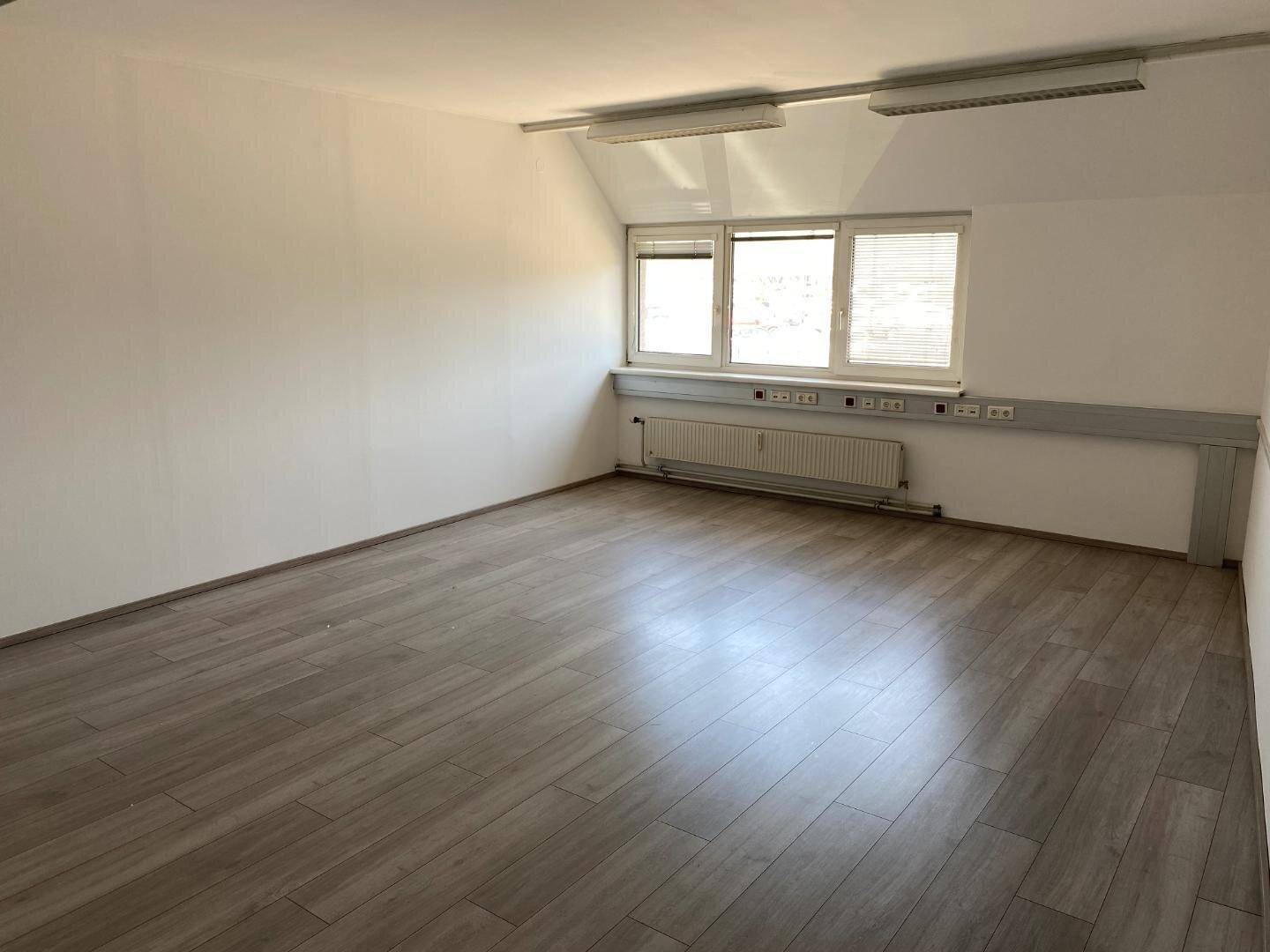 Leerer Büroraum mit großen Fenstern, die für eine helle Atmosphäre sorgen, und einem strapazierfähigen Laminatboden.