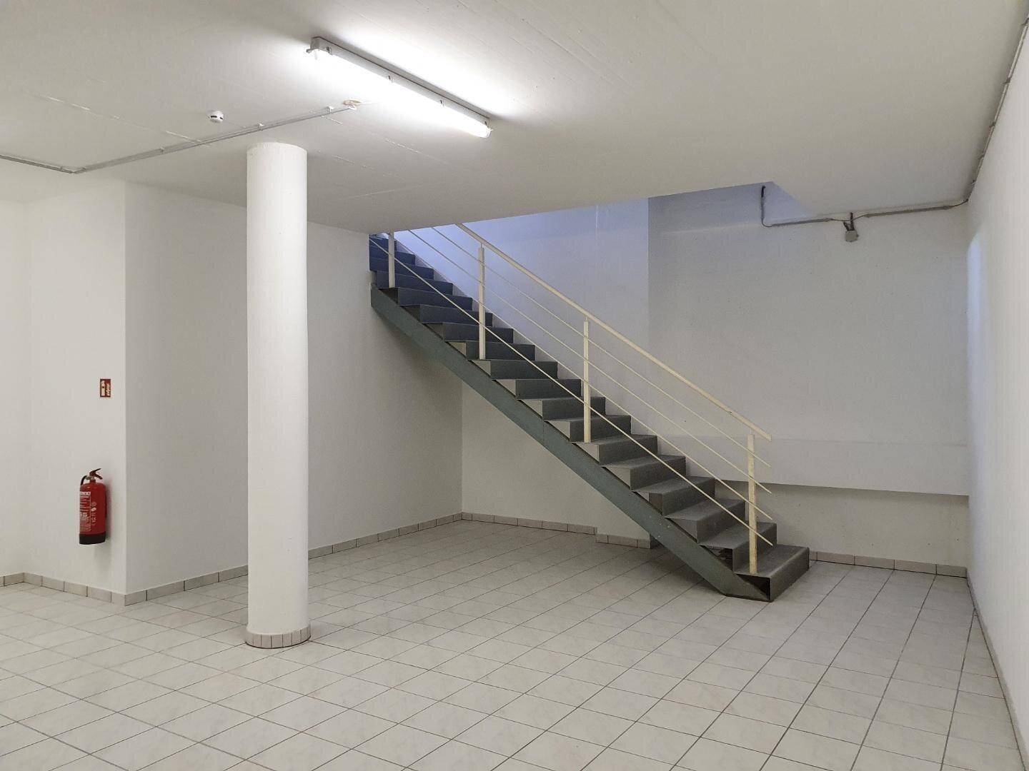 Heller Gewerberaum mit Treppe, tragender Säule und Fliesenboden, inklusive Feuerlöscher an der Wand.