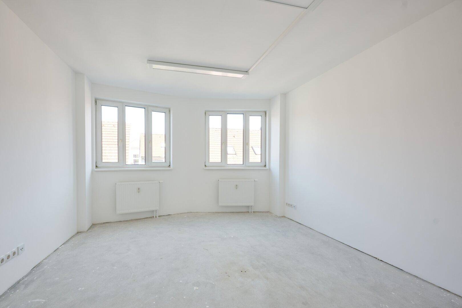 Heller Büroraum mit zwei Fenstern, weißen Wänden und einem unbehandelten Betonboden.