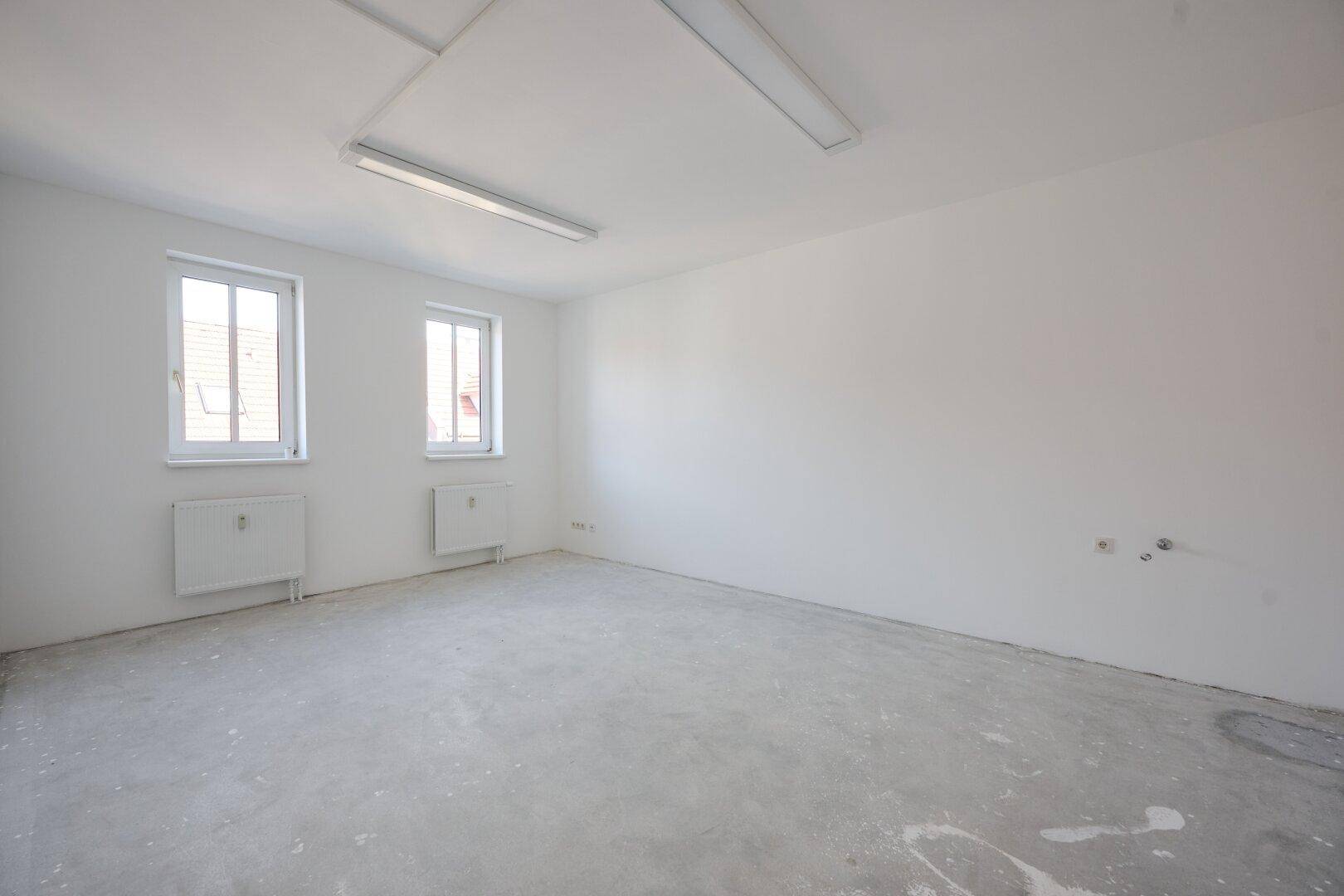Leerer Raum mit weißen Wänden, Betonboden und zwei Fenstern, die viel natürliches Licht bieten.