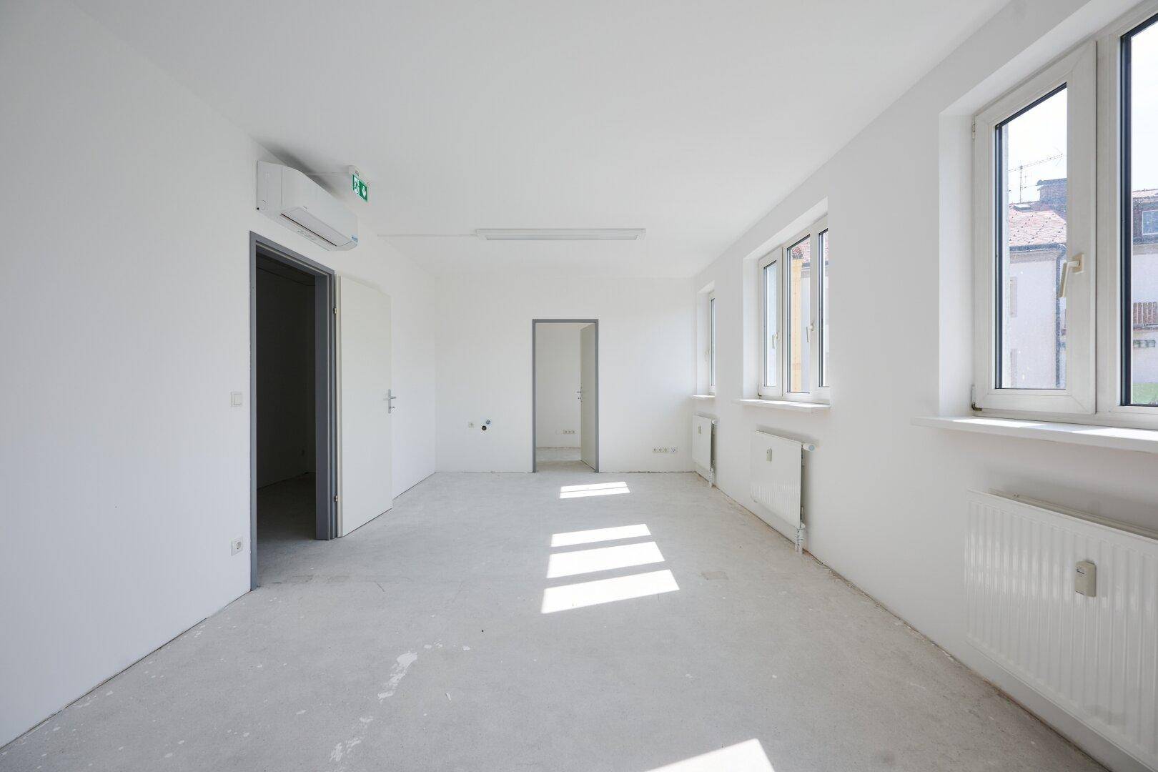 Geräumiger Büroraum mit Klimaanlage, großen Fenstern und Betonboden, durchflutet von Sonnenlicht.