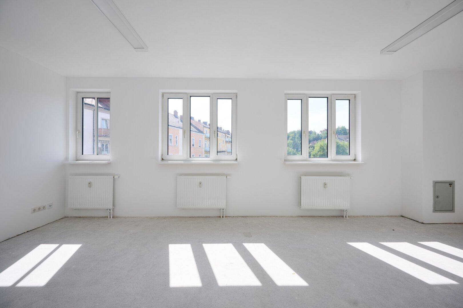 Heller Büroraum mit drei Fenstern, Betonboden und viel natürlichem Licht, das den Raum erhellt.