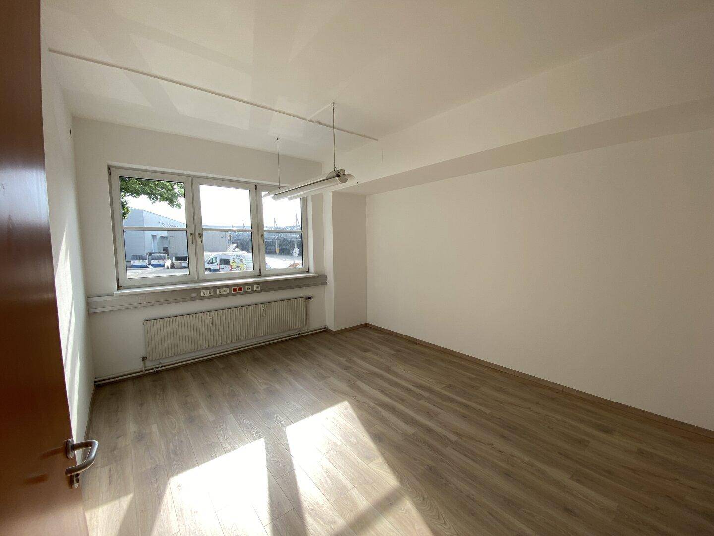 Heller Büroraum mit großem Fenster, Laminatboden und einem Heizkörper unter der Fensterbank, der für Wärme sorgt.