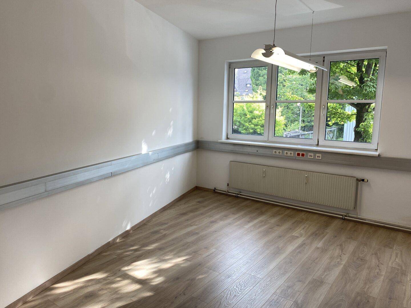 Heller Büroraum mit drei Fenstern, die einen Blick ins Grüne bieten, und einem Heizkörper unter der Fensterbank.