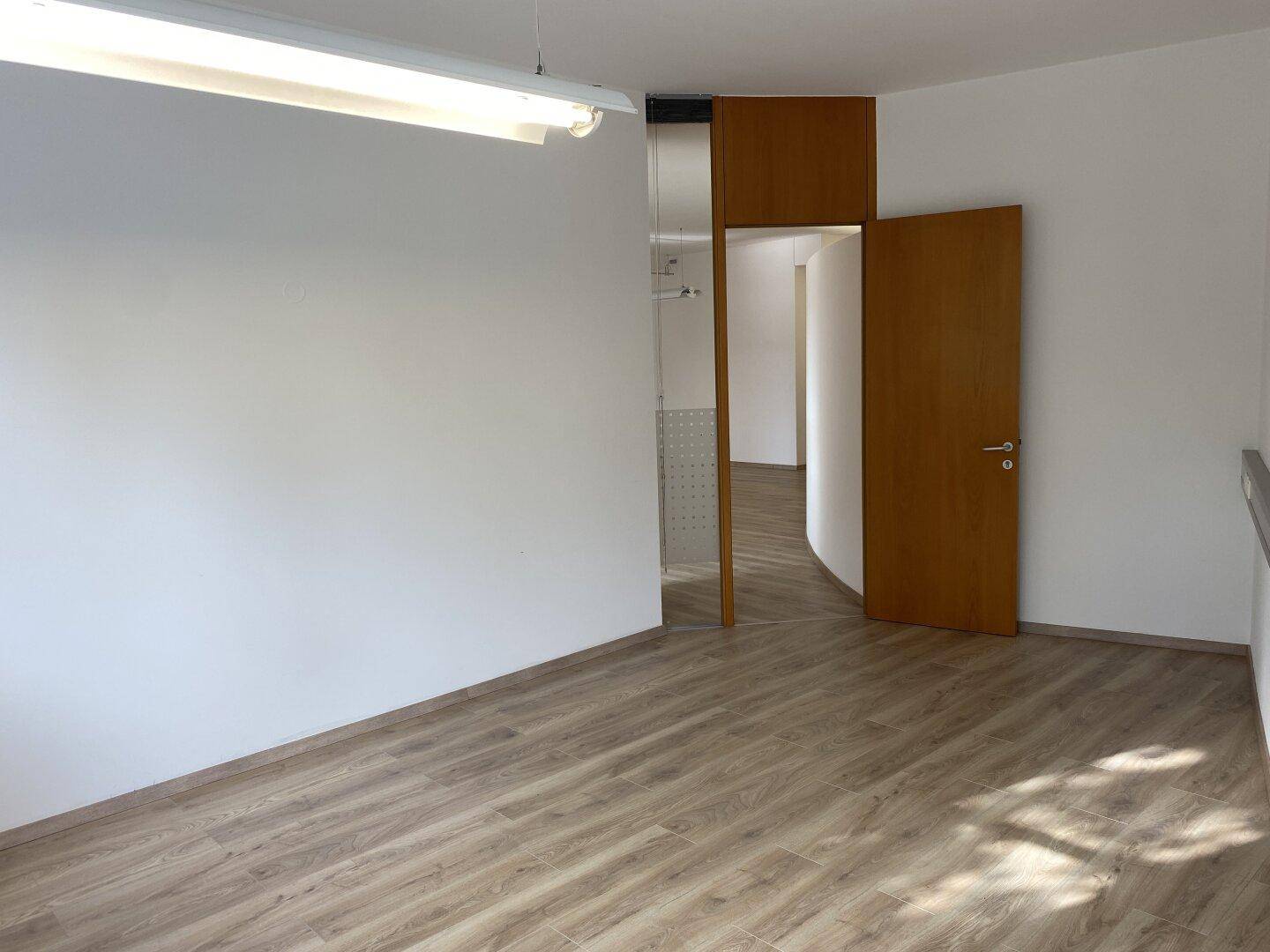Geräumiger Bürobereich mit Laminatboden, weißer Wand und einer Holztür, die zu einem weiteren Raum führt.