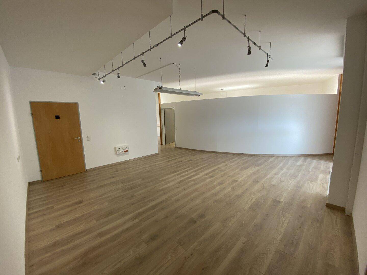 Heller Büroraum mit modernem Laminatboden, weiß gestrichenen Wänden und Schienenspots an der Decke für flexible Beleuchtung.