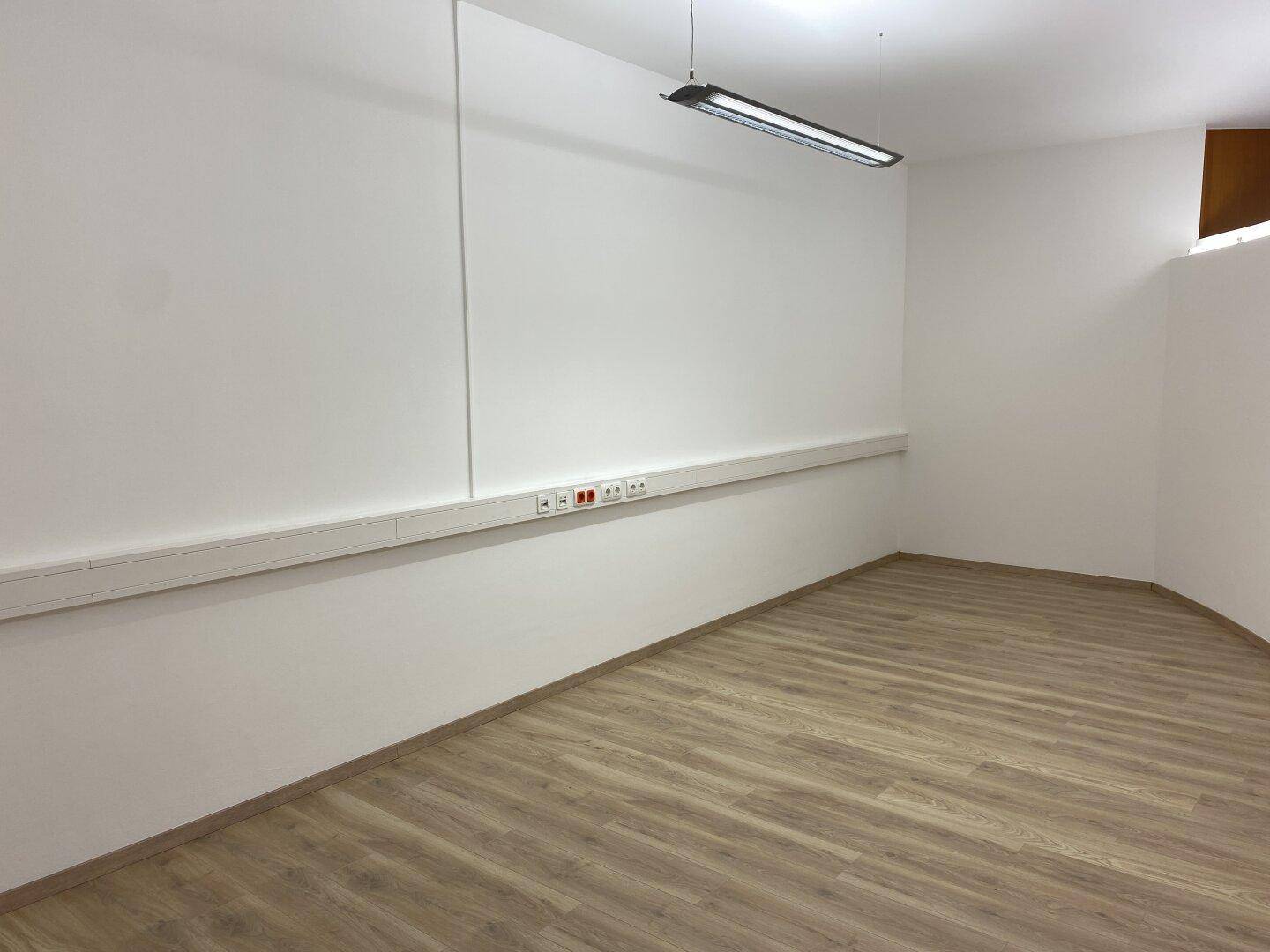 Leerer Büroraum mit Laminatboden, weißen Wänden und einer langen Steckdosenleiste entlang der Wand für Arbeitsplätze.