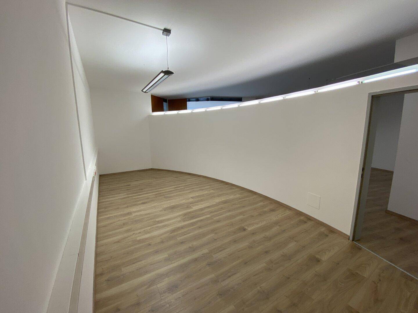 Heller Bürobereich mit geschwungener Wand, Laminatboden und indirekter Beleuchtung an der Decke, die eine angenehme Atmosphäre schafft.