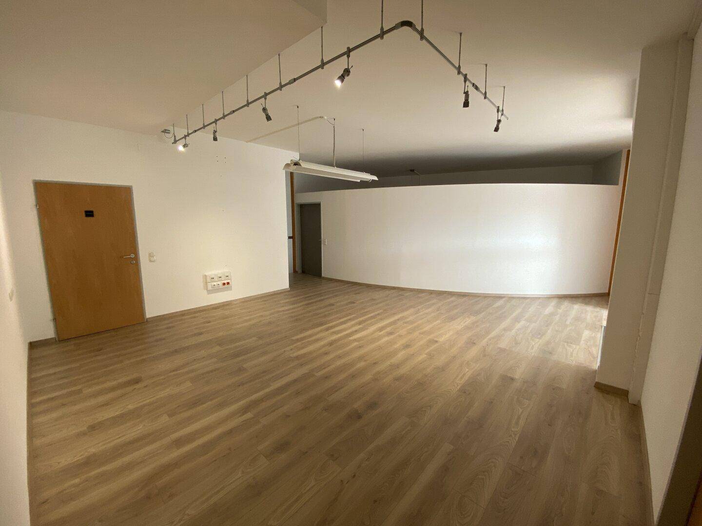 Geräumiger Büroraum mit hellem Laminatboden, weiß gestrichenen Wänden und Schienenspots an der Decke für flexible Beleuchtung.