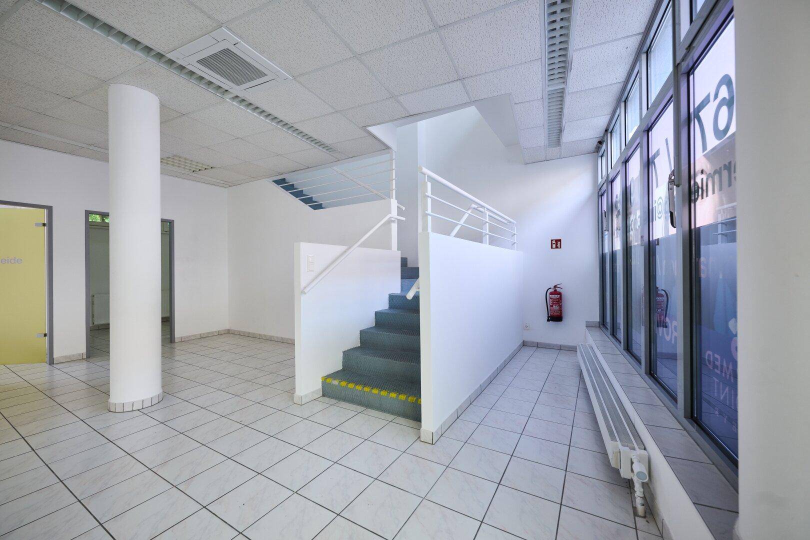 Treppenaufgang in einem hellen Gewerberaum mit großen Fenstern und Fliesenboden.