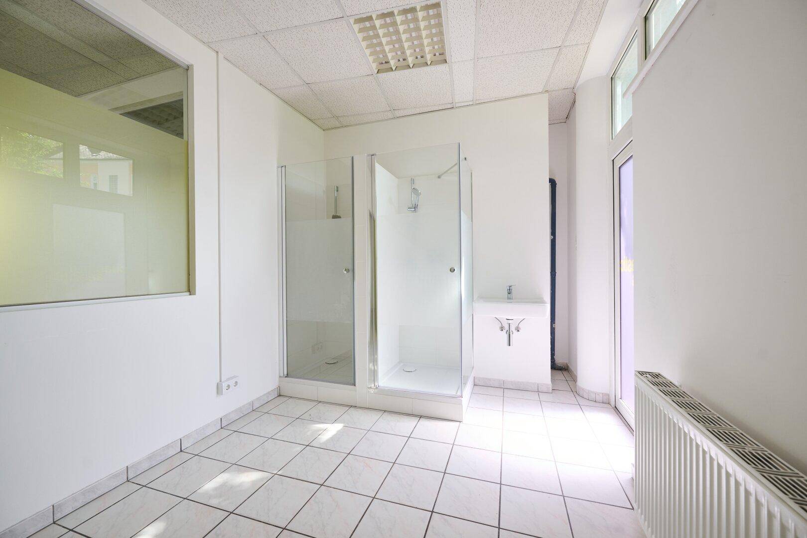 Raum mit zwei Duschkabinen, einem kleinen Waschbecken und weißen Fliesenboden.