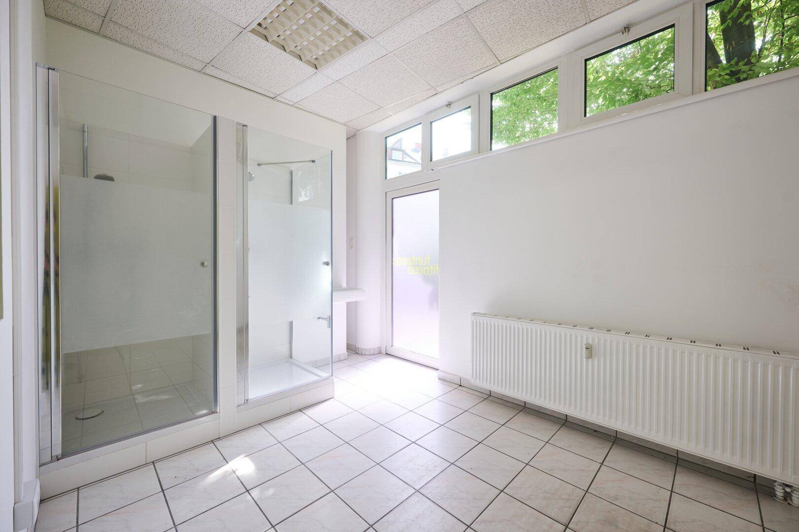 Raum mit zwei Duschkabinen, Heizkörper und hochgesetzten Fenstern für Tageslicht.