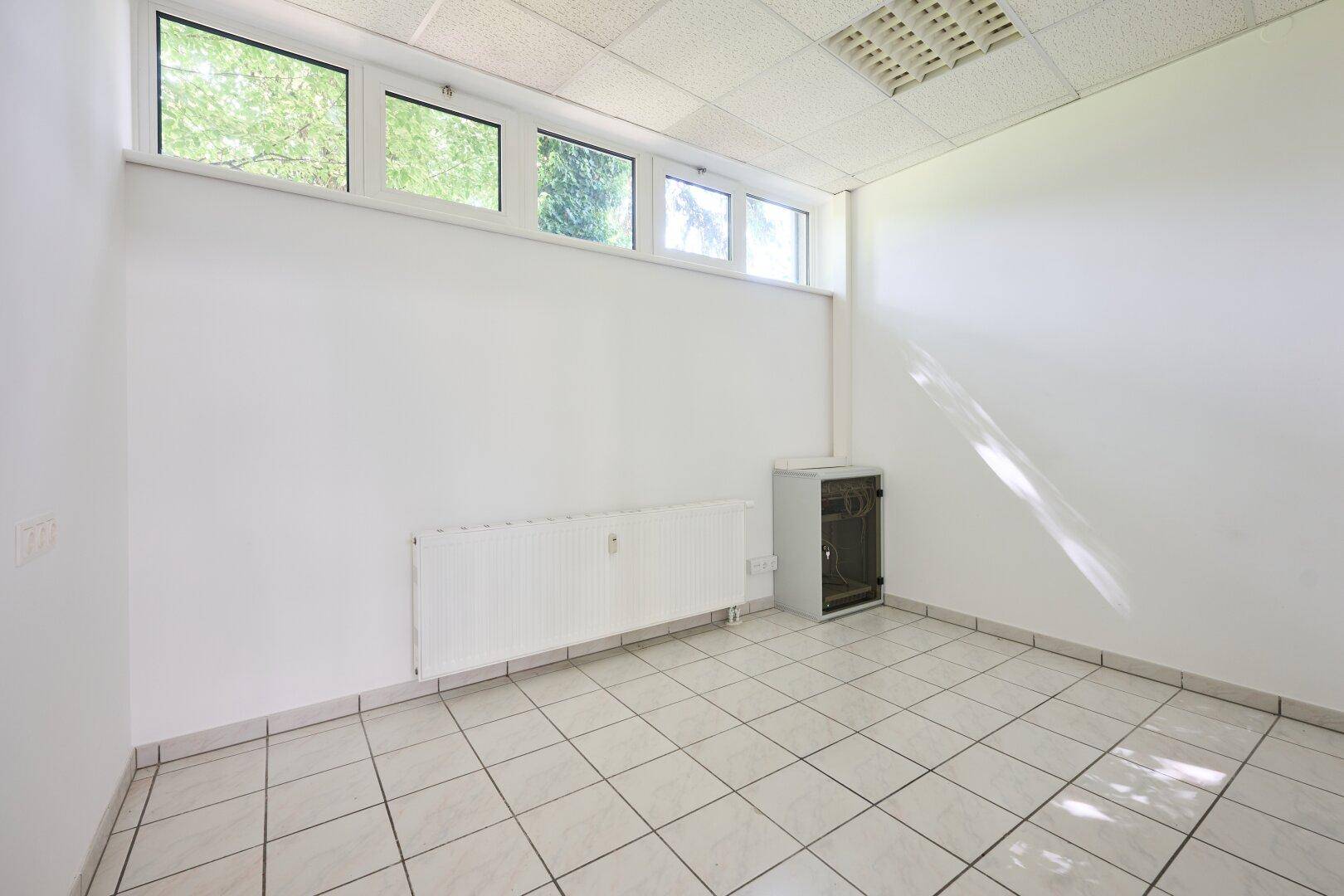 Leerer Raum mit weißen Wänden, Fliesenboden, Heizkörper und hochgesetzten Fenstern.
