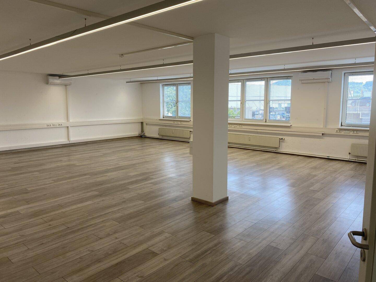 Geräumiger, leerer Büroraum mit großen Fenstern und Laminatboden, ideal für flexible Nutzung.