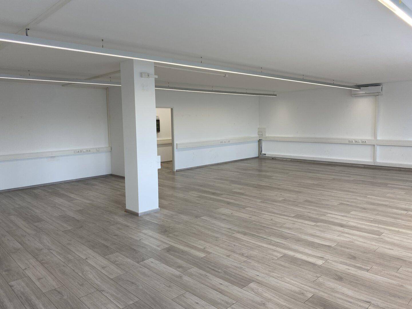 Heller, leerer Büroraum mit zentraler Säule, Laminatboden und modernen Deckenleuchten.
