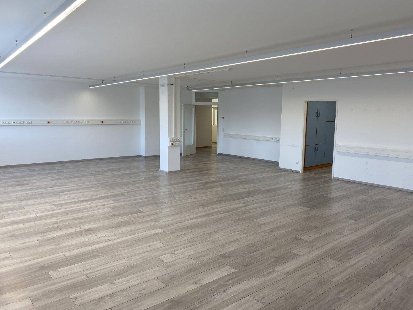 Großer, leerer Büroraum mit mehreren Türen, Laminatboden und durchgehenden Lichtleisten.