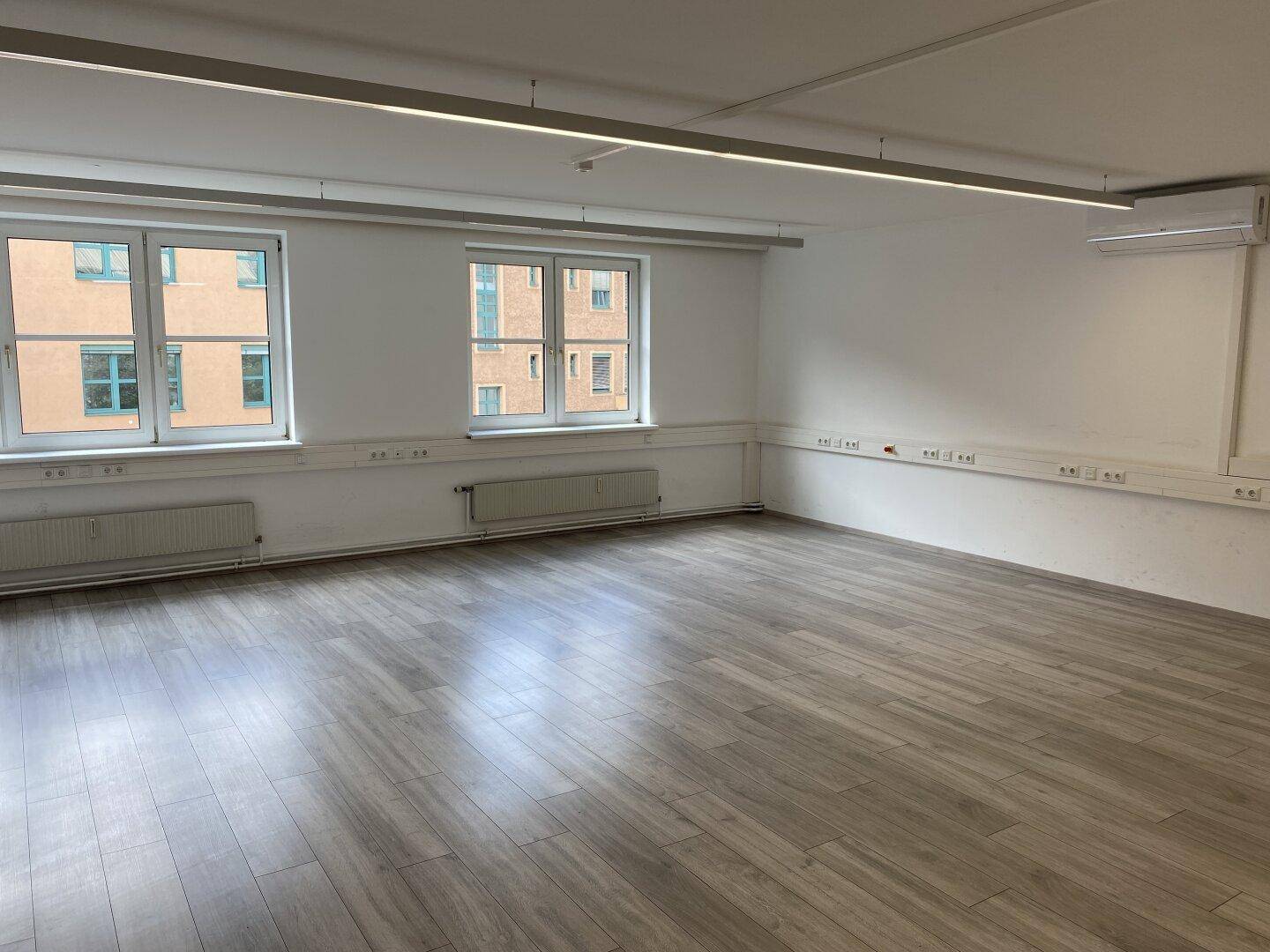 Leerer Büroraum mit zwei großen Fenstern, Laminatboden und weißen Wänden.