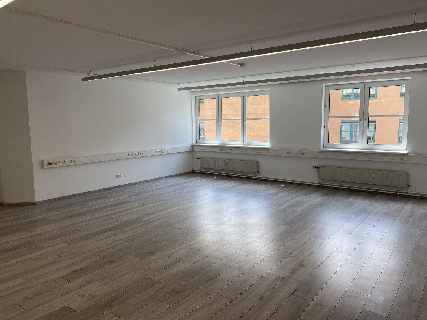 Heller, leerer Büroraum mit zwei Fenstern, Laminatboden und weißen Wänden.