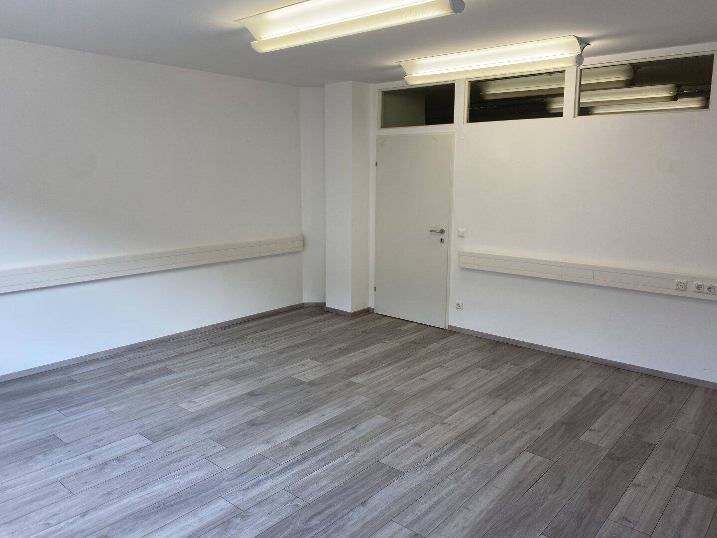 Geräumiger, leerer Büroraum mit Tür, Oberlichtern und Laminatboden.