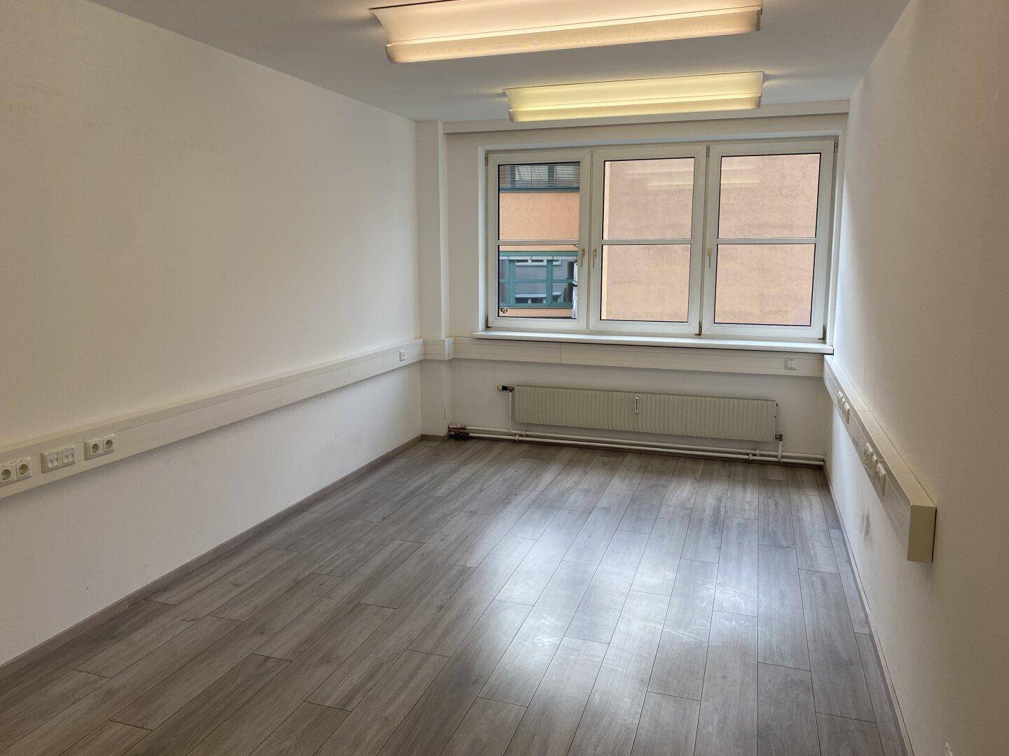 Leerer Büroraum mit drei Fenstern, Laminatboden und weißen Wänden, viel Tageslicht.
