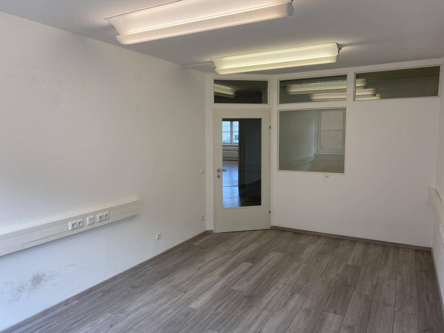 Heller, leerer Büroraum mit Glastür, Oberlichtern und Laminatboden.