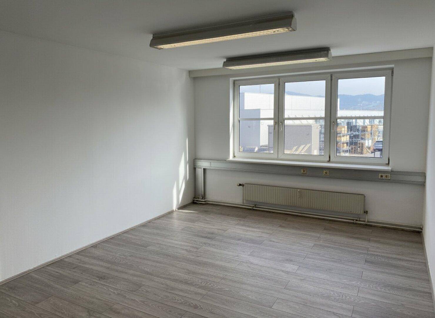 Leerer Büroraum mit großen Fenstern, Laminatboden und Blick auf die Industrieumgebung.