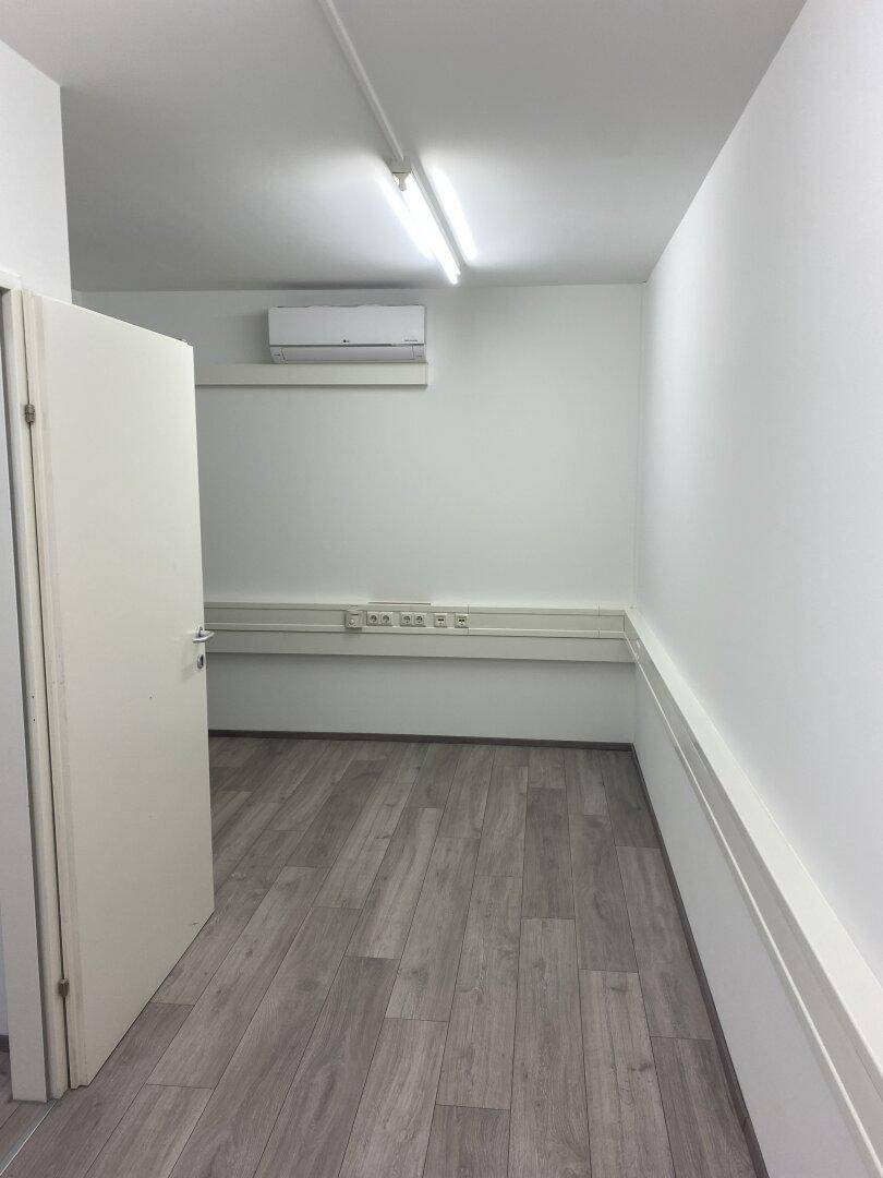Schmaler Büroraum mit Laminatboden, weiß gestrichenen Wänden und Klimaanlage, ideal für Einzelarbeitsplätze.