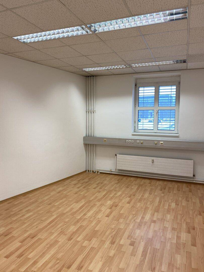 Helles Büro mit Laminatboden, Deckenleuchten und einem Fenster mit Jalousien.