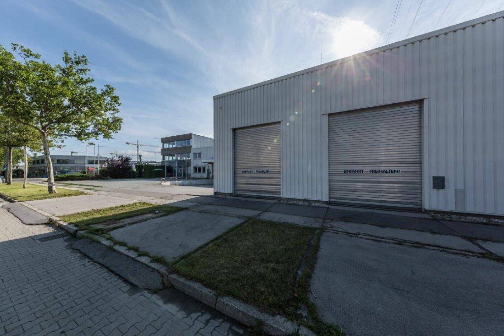 Außenseite eines Industriegebäudes mit zwei großen Rolltoren und einem sonnigen Vorplatz.
