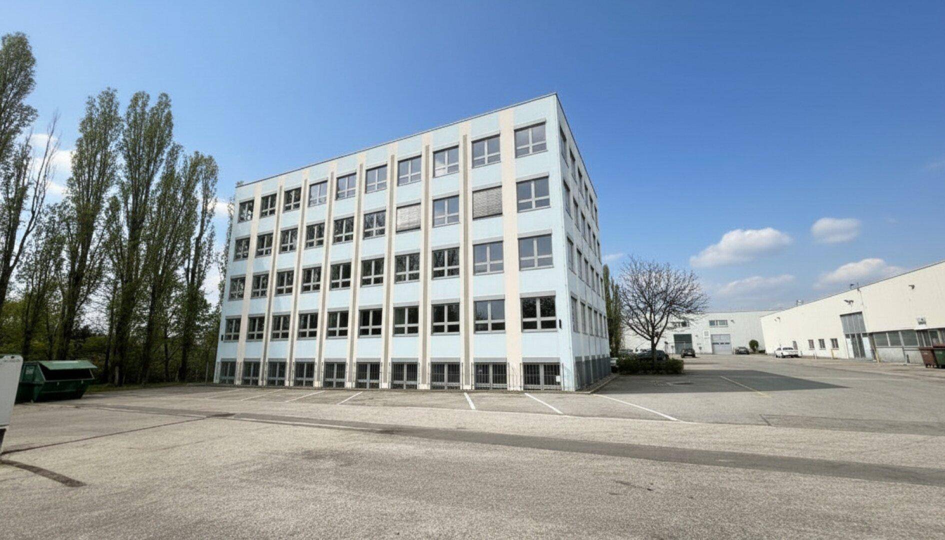 Weitläufige Ansicht des modernen Bürogebäudes mit umliegenden Parkplätzen und Bäumen unter klarem Himmel.