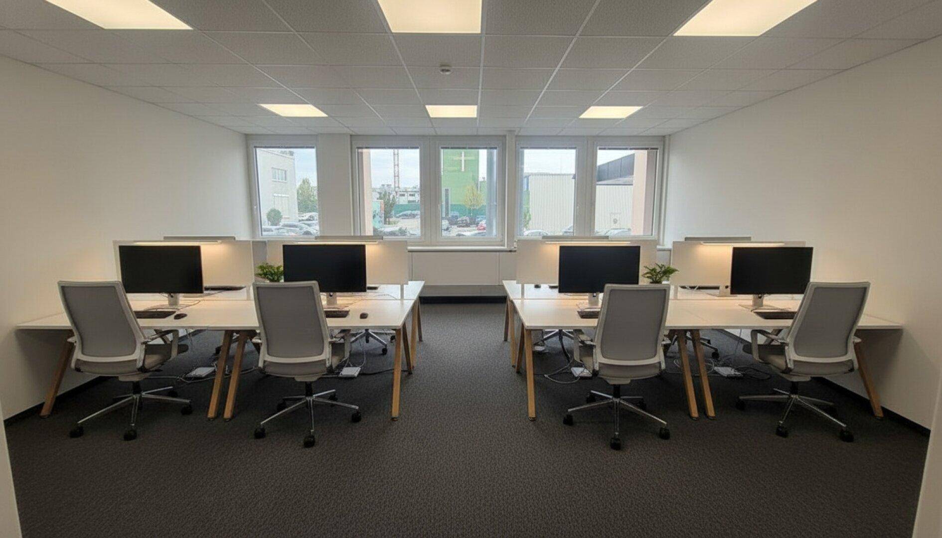 Heller Großraumbüro mit mehreren Arbeitsplätzen, ergonomischen Stühlen und großen Fenstern für Tageslicht.