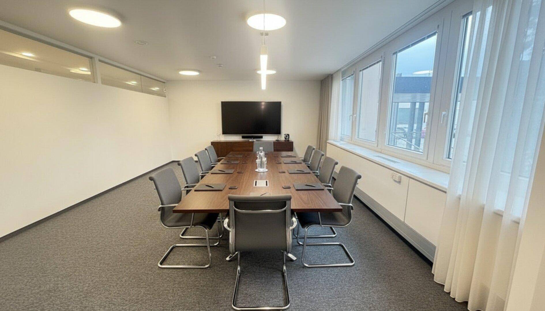 Großer Konferenzraum mit langem Holztisch, bequemen Stühlen und einem großen Bildschirm für Präsentationen.
