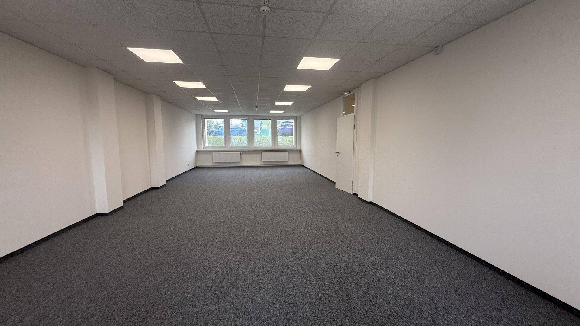 Geräumiger, leerer Büroraum mit Teppichboden und vielen Fenstern für natürliches Licht.