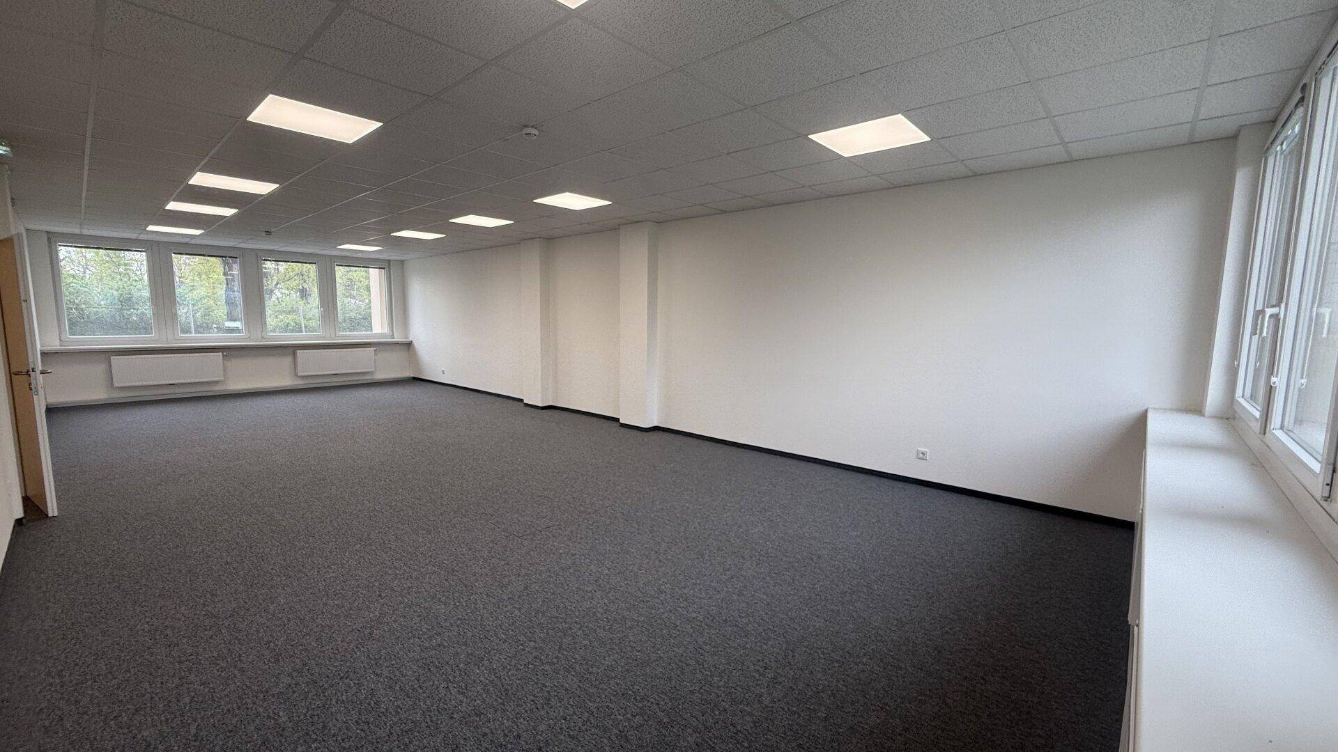 Großer, unmöblierter Büroraum mit Teppichboden, weißen Wänden und vielen Fenstern.